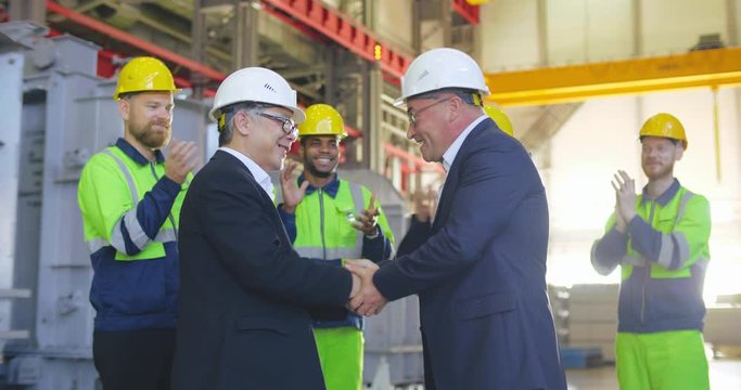 4K Businessmen shake hands on the factory after successful deal. Group of international partners congrats each other with success. Workmen applause on background