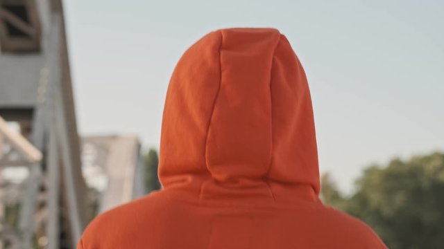 Back view of an athletic person with a hood on his head wearing orange hoodie is running on the bridge in the morning