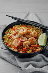 Shrimp fried rice with vegetables, green onions and lime in a black pottery bowl on a fabric tablecloth. Traditional Thai meal with sesame on a white table. Shot with space for text.