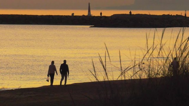 Older couple walking on beach together at dawn, silhouette against calm sea, slow motion
