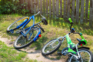 Fototapeta premium Children's and adult bicycles lay in the grass on the side of the road near a rural fence. Country trips on the weekend
