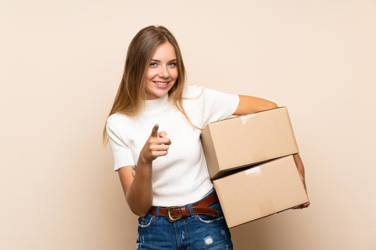 Young blonde woman over isolated background holding a box to move it to another site while pointing to the front