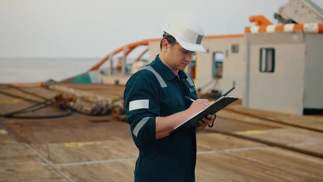Marine chief officer or chief mate on deck of ship or vessel. He fills up ahts vessel checklist. Ship routine paperwork. He holds VHF walkie-talkie radio in hands.