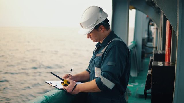 Marine chief officer or chief mate on deck of ship or vessel. He fills up ahts vessel checklist. Ship routine paperwork. He holds VHF walkie-talkie radio in hands.