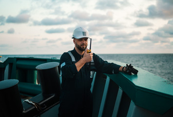 Obraz premium Marine Deck Officer or Chief mate on deck of offshore vessel or ship , wearing PPE personal protective equipment - helmet, coverall. He holds VHF walkie-talkie radio in hands.
