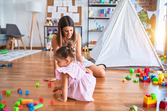 Young beautiful teacher and toddler sitting on the floor playing with building blocks toy at kindergarten