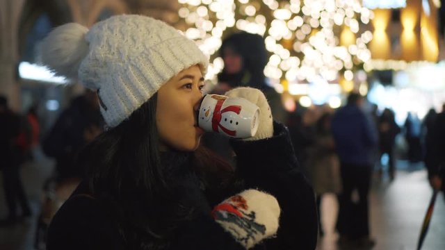 Pretty young girl in white hat is drinking hot cocktail outside at Christmas. Decorations for Christmas in european city, New Year tree with lights and garlands