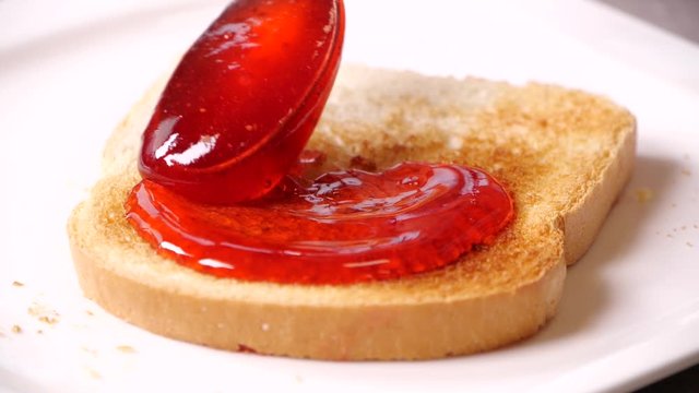 Spreading of delicious strawberry jam onto crispy toast, closeup. Slow motion effect