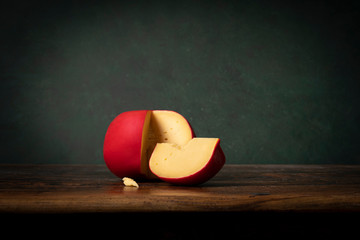 A head of Edam cheese with a red crust lying on a brouwn wooden table with a green background....