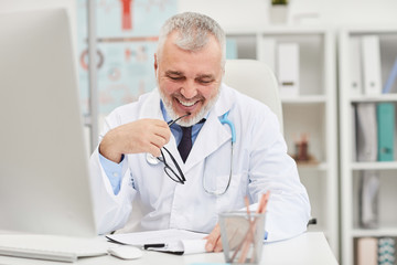 Fototapeta premium Smiling mature doctor sitting at his workplace in front of computer and examining the document