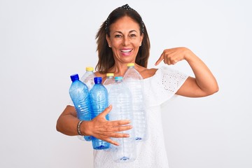 Middle age beautiful woman recycling plastic bottles standing over isolated white background with surprise face pointing finger to himself