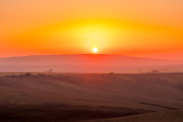 Obraz na płótnie Canvas The sun caught during the last moment before setting beyond the horizon with view of a field covered with fog and the farms and fields undulating in the region of South Moravia is also called Tuscany