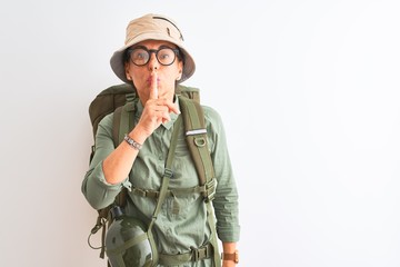 Middle age hiker woman wearing backpack canteen hat glasses over isolated white background asking to be quiet with finger on lips. Silence and secret concept.