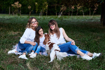 Two young blond women and one brunette girl, wearing jeans and white t-shirts, sitting on green blanket in park, playing with cavalier king charles spaniel, smiling, laughing. Leisure time in summer.