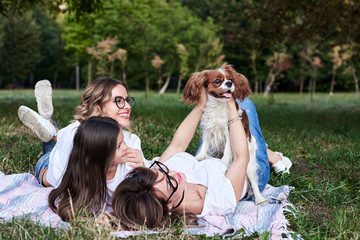 Two young blond women and one brunette girl, wearing white t-shirts, lying on mexican blanket on green grass in park, holding cavalier king charles spaniel, smiling, laughing. Picnic in summertime.