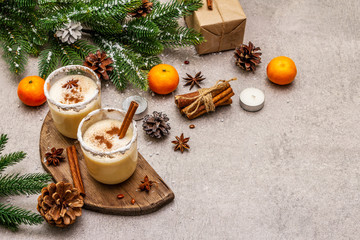 Eggnog with cinnamon and nutmeg for Christmas and winter holidays. Homemade beverage in glasses with spicy rim. Tangerines, candles, gift. Stone concrete background