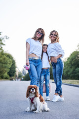 Three young girls, wearing blue jeans and white t-shirts, walking with small dog in city park in summer. Family fun leisure time with cavalier king charles spaniel. Pretty women posing for picture.