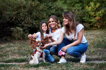 Two young blond women and one brunette girl, wearing casual clothes, kneeling on green grass in park, holding cavalier king charles spaniel, smiling, laughing. Family walk with little dog in forest.