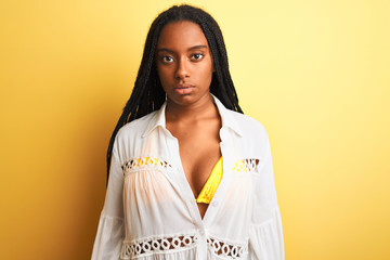 Fototapeta premium African american woman on vacation wearing bikini and shirt over isolated yellow background with serious expression on face. Simple and natural looking at the camera.