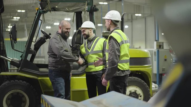 Young Caucasian men standing at the background of tow tractor and talking as driver getting out the cabin and shaking their hands. Workers in manufacture plant. Professional, working, loader, employee