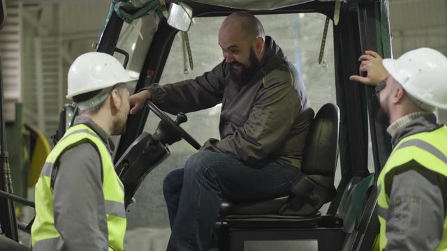 Portrait of bearded Caucasian tow tractor driver sitting in cabin and talking to his colleagues. Workers in vests and helmets having break at production site. Manufacture, loader, production.