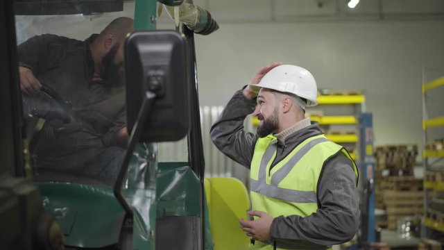 Profile portrait of young bearded Caucasian man putting on helmet and talking to his colleague in tow tractor. Workers on manufacture plant. Manufacturing, production, professionals. occupation.