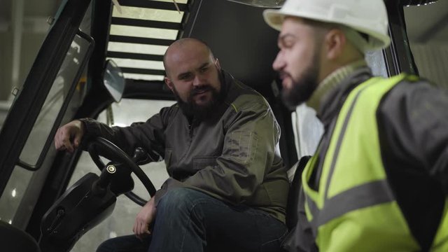 Blurred bearded Caucasian man in helmet and vest talking to tow tractor driver. Worker sitting in cabin and listening to colleague. Employees discussing schedule at warehouse. Manufacture, industry.