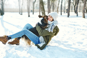 Young happy woman in woolen hat and long warm scarf hugs her pet in snowy winter park at frozzy sunny day. Happy time together, cute dog Welsh Corgi Pembroke, winter holodays.