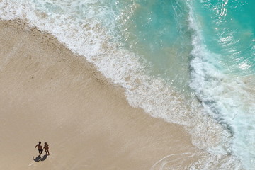 Top view of a couple walking on the beautiful beach with huge blue and turquoise waves in the sea. Travel, holidays inspiration, lifestyle, weekend getaway concept
