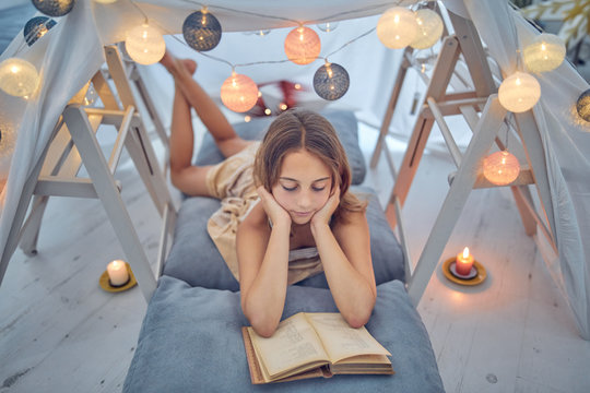 Little 10 Year Old Girl Reading Classic Book Under Her Home-made Tent Inside The Living Room.