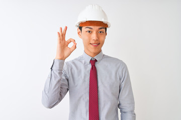 Chinese architect man wearing tie and helmet standing over isolated white background smiling positive doing ok sign with hand and fingers. Successful expression.