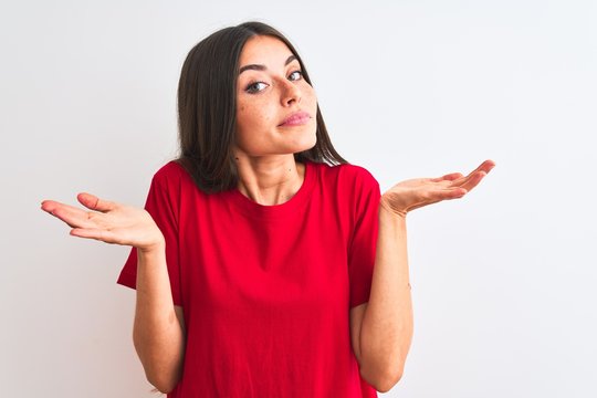 Young beautiful woman wearing red casual t-shirt standing over isolated white background clueless and confused expression with arms and hands raised. Doubt concept.