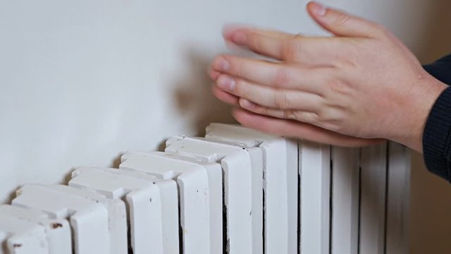 A man in a hat warms his hands on a heating radiator near the wall. Cold in the apartment, poor heating system