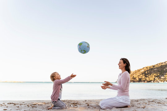 Happy little girl sitting with her mother on the beach playing with Earth beach ball