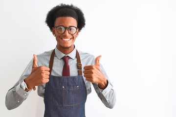 Young african american shopkeeper man wearing apron glasses over isolated white background success sign doing positive gesture with hand, thumbs up smiling and happy. Cheerful expression