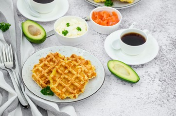 Cheese crispy waffles with cream cheese, smoked salmon and avocado for breakfast with a cup of coffee on a light stone background.