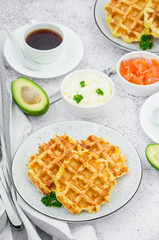 Cheese crispy waffles with cream cheese, smoked salmon and avocado for breakfast with a cup of coffee on a light stone background.