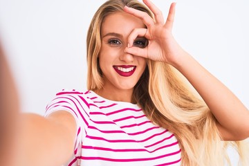 Beautiful woman wearing striped t-shirt make selfie by camera over isolated white background with happy face smiling doing ok sign with hand on eye looking through fingers