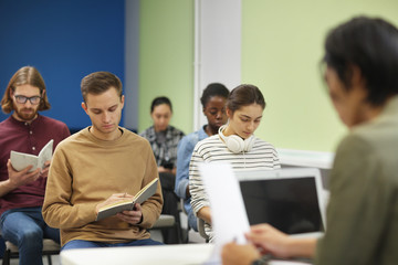 Multiethnic group of young people sitting and writing in notebooks while teacher reading a lecture