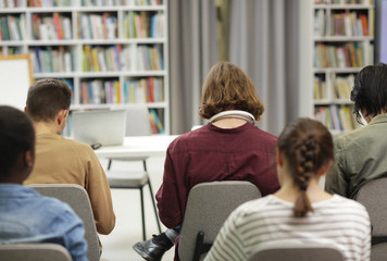 Rear view of group of young people sitting on chairs and working in the library