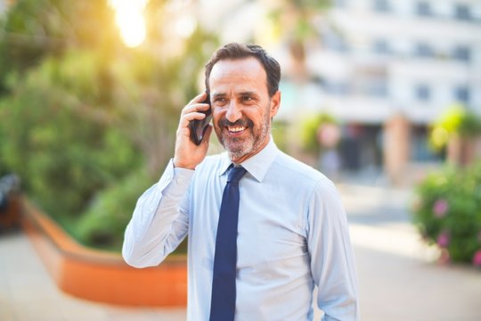 Middle age handsome businessman standing on the street talking on the smartphone smiling