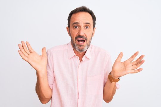 Middle age handsome man wearing casual pink shirt standing over isolated white background clueless and confused expression with arms and hands raised. Doubt concept.