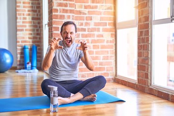 Middle age handsome sportman sitting on mat doing stretching yoga exercise at gym smiling funny doing claw gesture as cat, aggressive and sexy expression