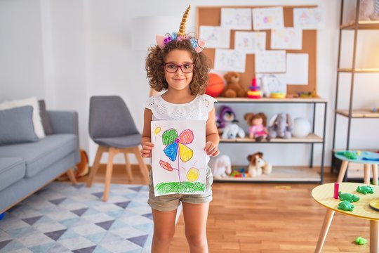 Beautiful toddler wearing glasses and unicorn diadem standing holding draw smiling at kindergarten