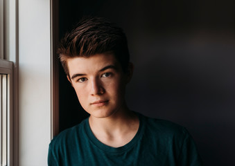 Portrait of caucasian teenage boy beside window with dark background.