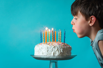 Boy blowing out candles on a birthday cake against blue background.