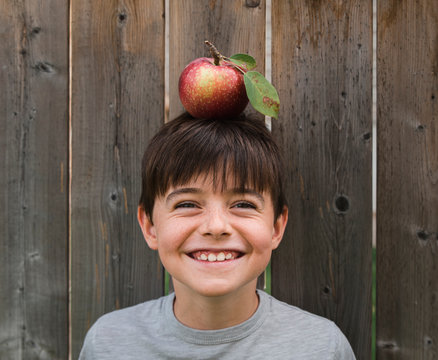 Boy Smiling While Balancing An Apple On His Head Against Wooden Fence