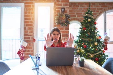 Beautiful woman sitting at the table working with laptop at home around christmas tree doing stop gesture with hands palms, angry and frustration expression