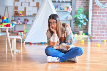 Young beautiful teacher woman wearing sweater and glasses sitting on the floor at kindergarten thinking looking tired and bored with depression problems with crossed arms.