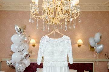 Beautiful wedding dress hanging on hanger on luxury chandelier in hotel room, copy space, close up. Bridal morning preparations